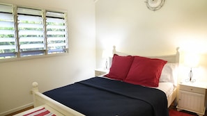 A bedroom with a bed, white nightstands, a window with blinds, and a wall-mounted light fixture.