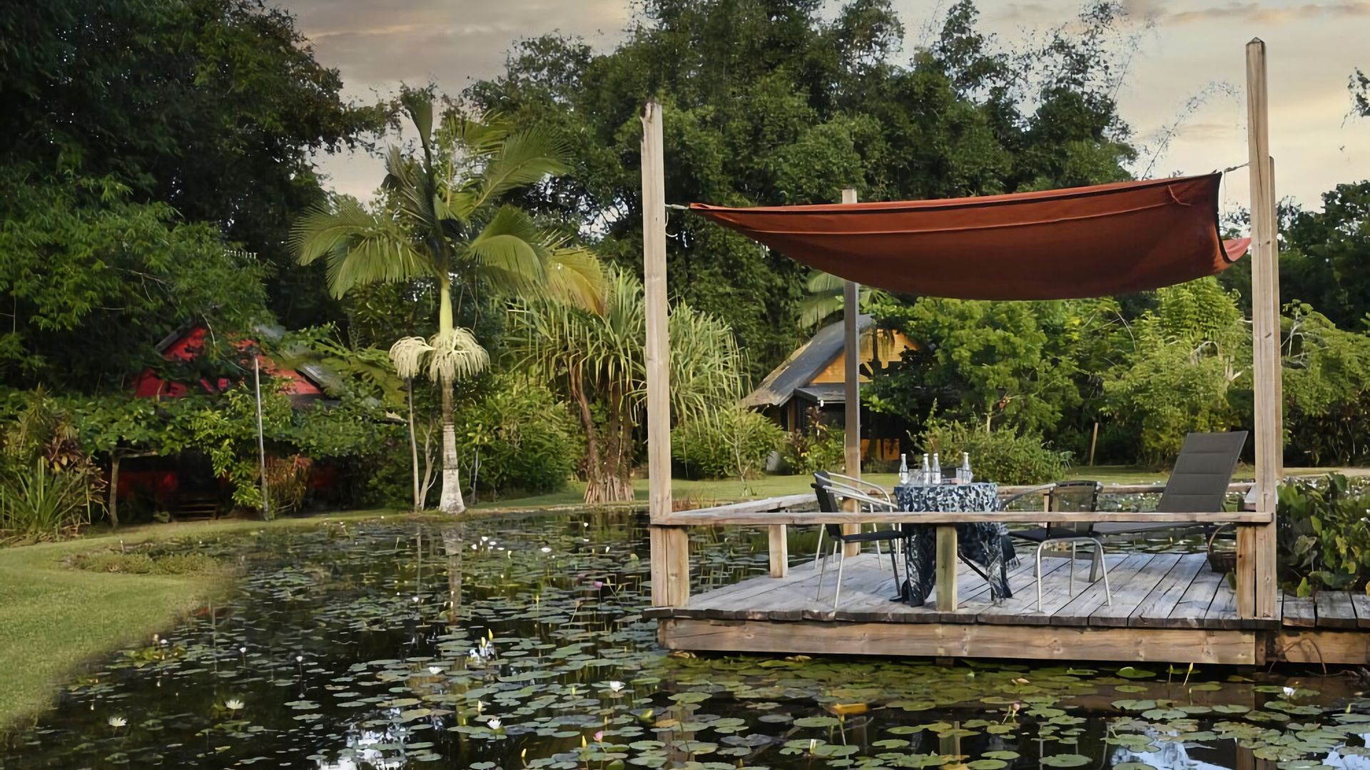 A wooden dock with a table and chairs, surrounded by a pond with lily pads.