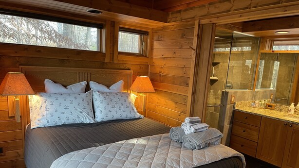 A wooden cabin room with a bed, two bedside lamps, a bathroom with a mirror, and a window with a view of trees. - Cabin amongst the trees (Waxhaw)