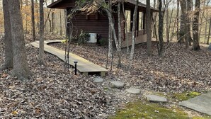 A rustic cabin with a wooden deck and a small porch, surrounded by trees and covered in fallen leaves. - Cabin amongst the trees (Waxhaw)