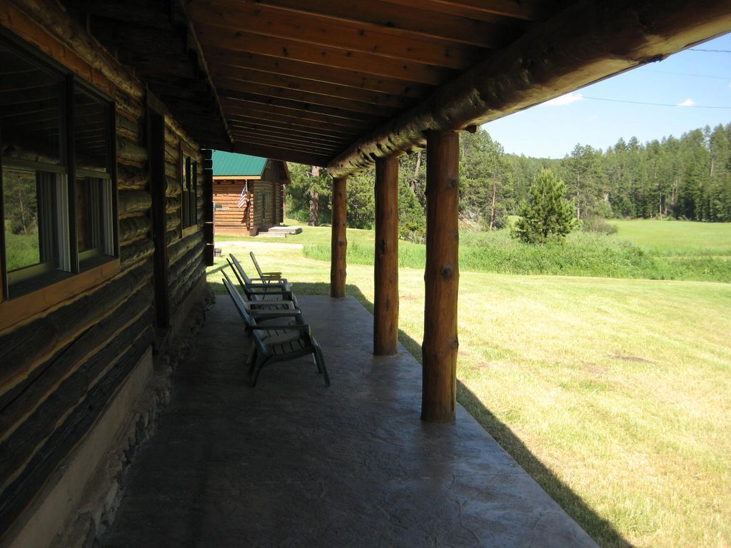 A log cabin with a covered porch, two chairs, and a view of a grassy field and forest.