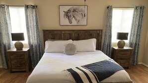 Una cama grande con cabecera de madera, dos mesitas de noche con lámparas, cortinas estampadas y un cuadro colgado en la pared.