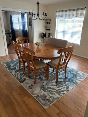 Comedor con mesa de madera, cuatro sillas, una lámpara de araña y alfombra con estampado floral.