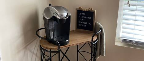 A coffee station with a kettle, coffee maker, and mugs on shelves, a sign with a coffee quote, and a window with blinds.