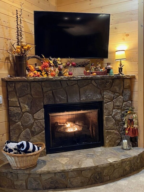 A stone fireplace with a lit fire, a television mounted above it, and a basket with clothes beside the fireplace. - Riverfront Cabin w/ Private Dock, Putting Green, Game Room! 3.5mi 2 Blue Ridge (Blue Ridge)