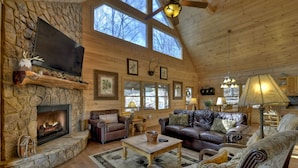 A cozy log cabin interior with a stone fireplace, leather sofas, and a large TV mounted on the wall. - Riverfront Cabin w/ Private Dock, Putting Green, Game Room!  3.5mi 2 Blue Ridge (Blue Ridge)