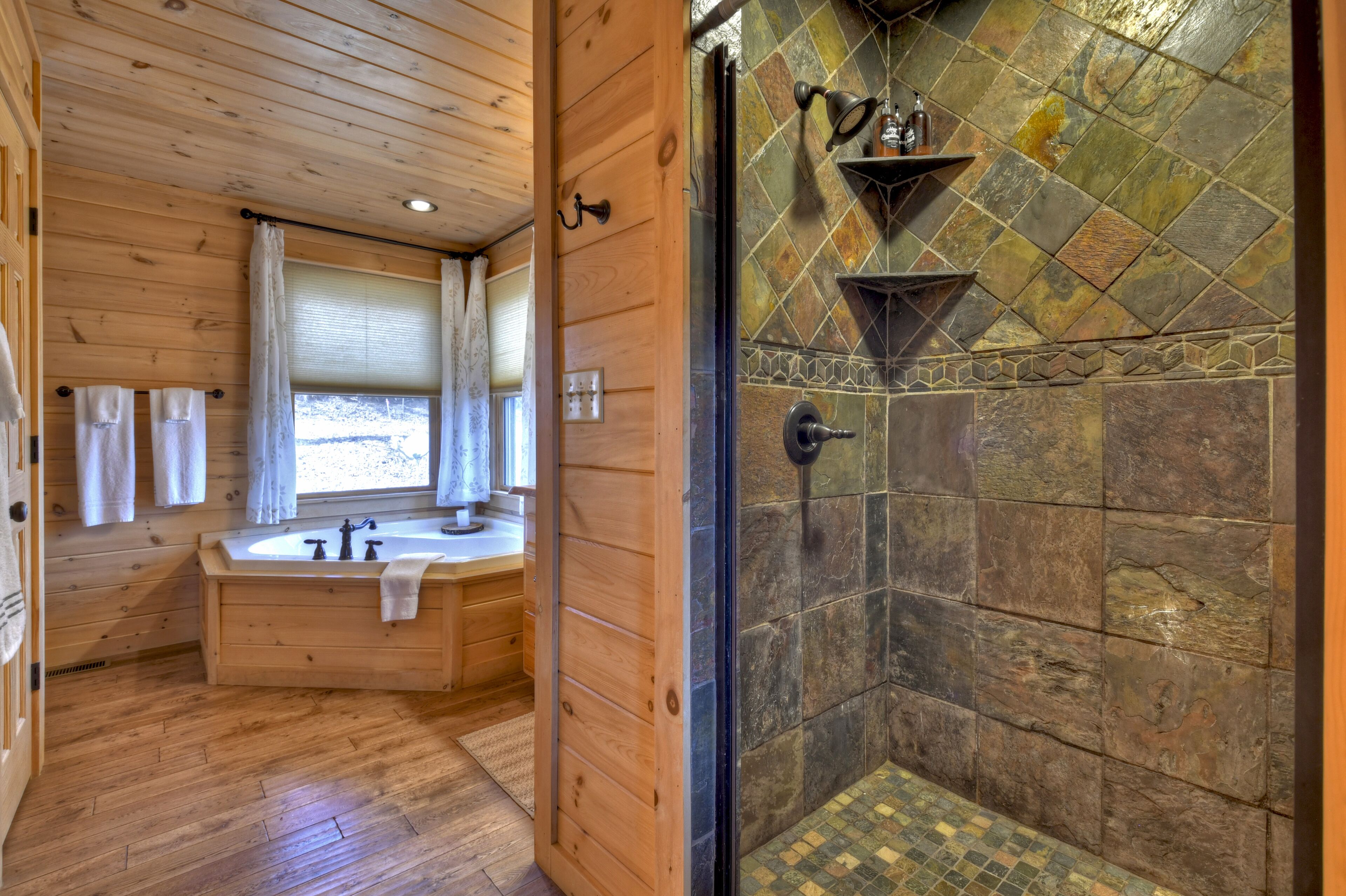 A wooden bathroom with a bathtub, a shower area with stone tiles, and a window with curtains.