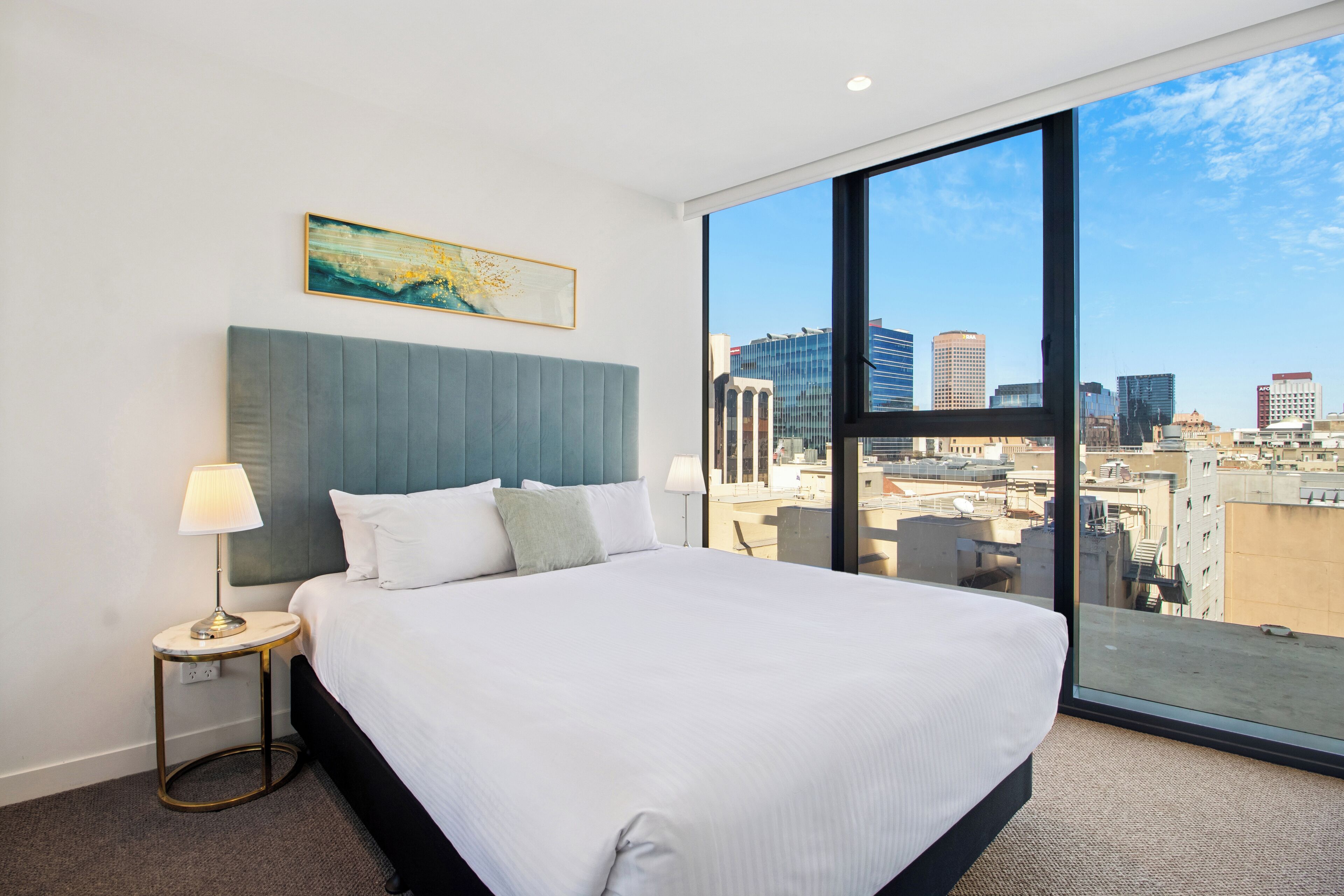 Ein Hotelzimmer mit einem großen Bett, einem runden Beistelltisch, einer Lampe, einem Gemälde und Blick auf die Stadt durch ein großes Fenster.
