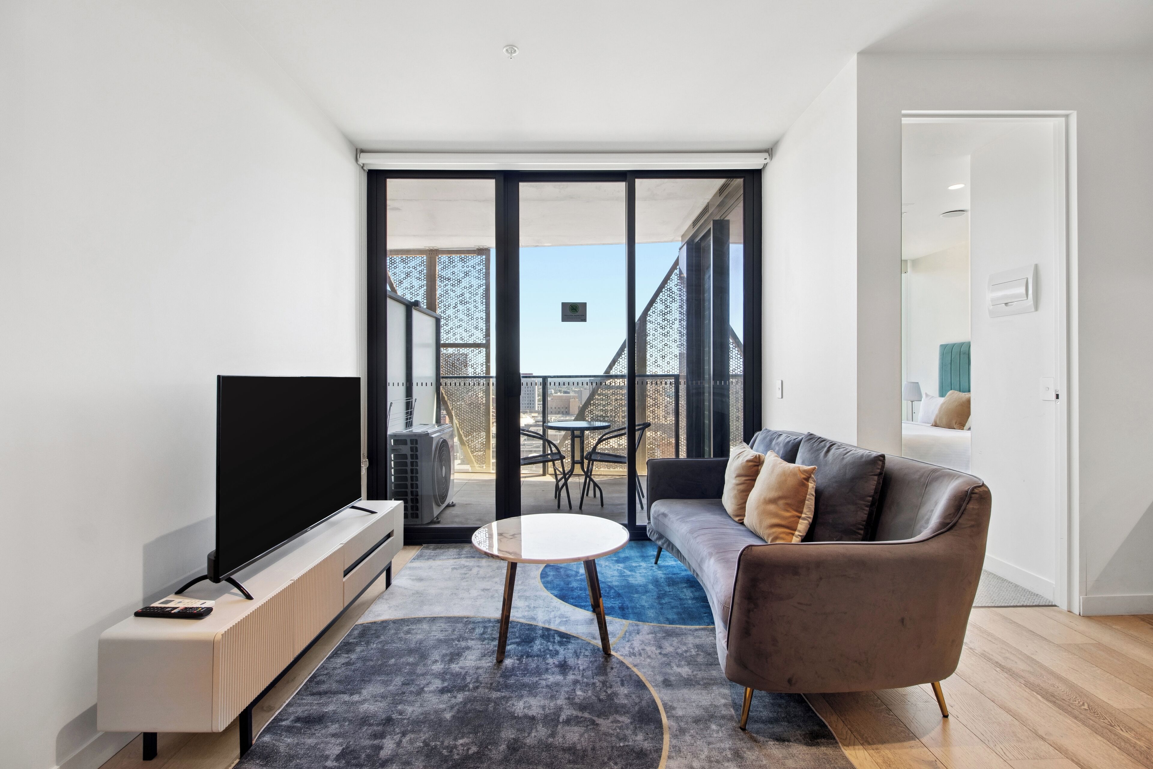 A modern living room with a flat-screen TV, a sofa, a round coffee table, and a balcony with a view.