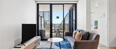 A modern living room with a flat-screen TV, a sofa, a round coffee table, and a balcony with a view.