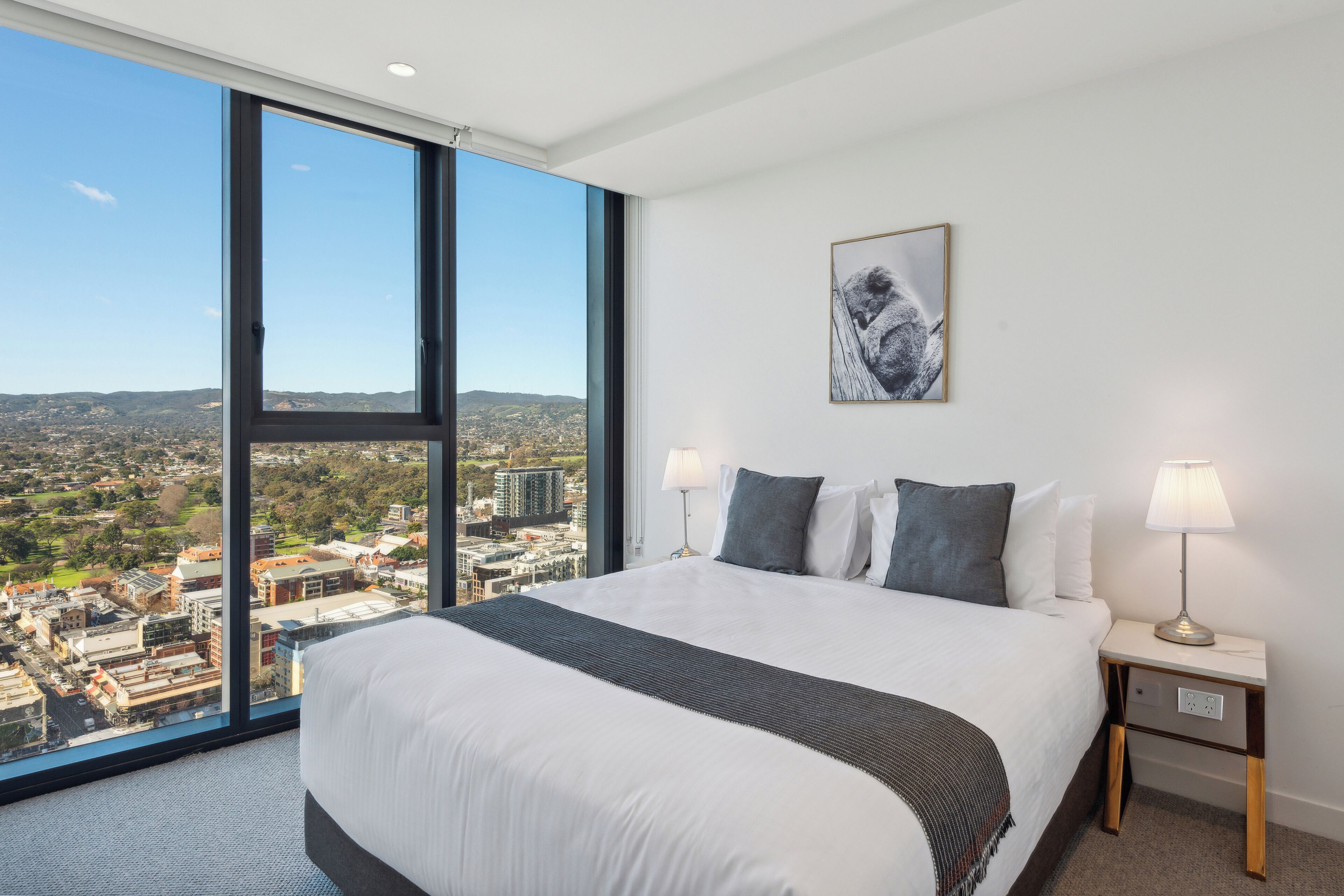 A hotel room with a large bed, two bedside tables, a nightstand, a lamp, a framed artwork, and a city view.