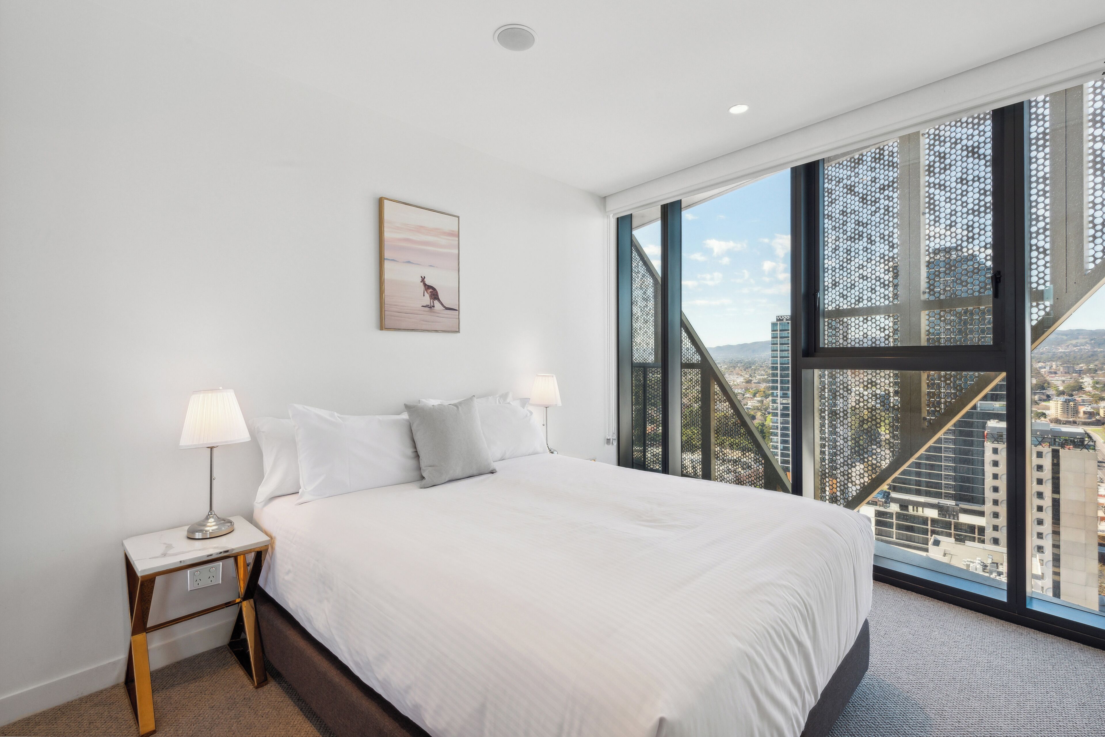 A modern hotel room with a large bed, two bedside lamps, a city view through a large window, and a framed picture on the wall.