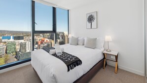 Una habitación de hotel moderna con una cama grande, vistas a la ciudad, una obra de arte enmarcadas y una mesita de noche con lámpara.