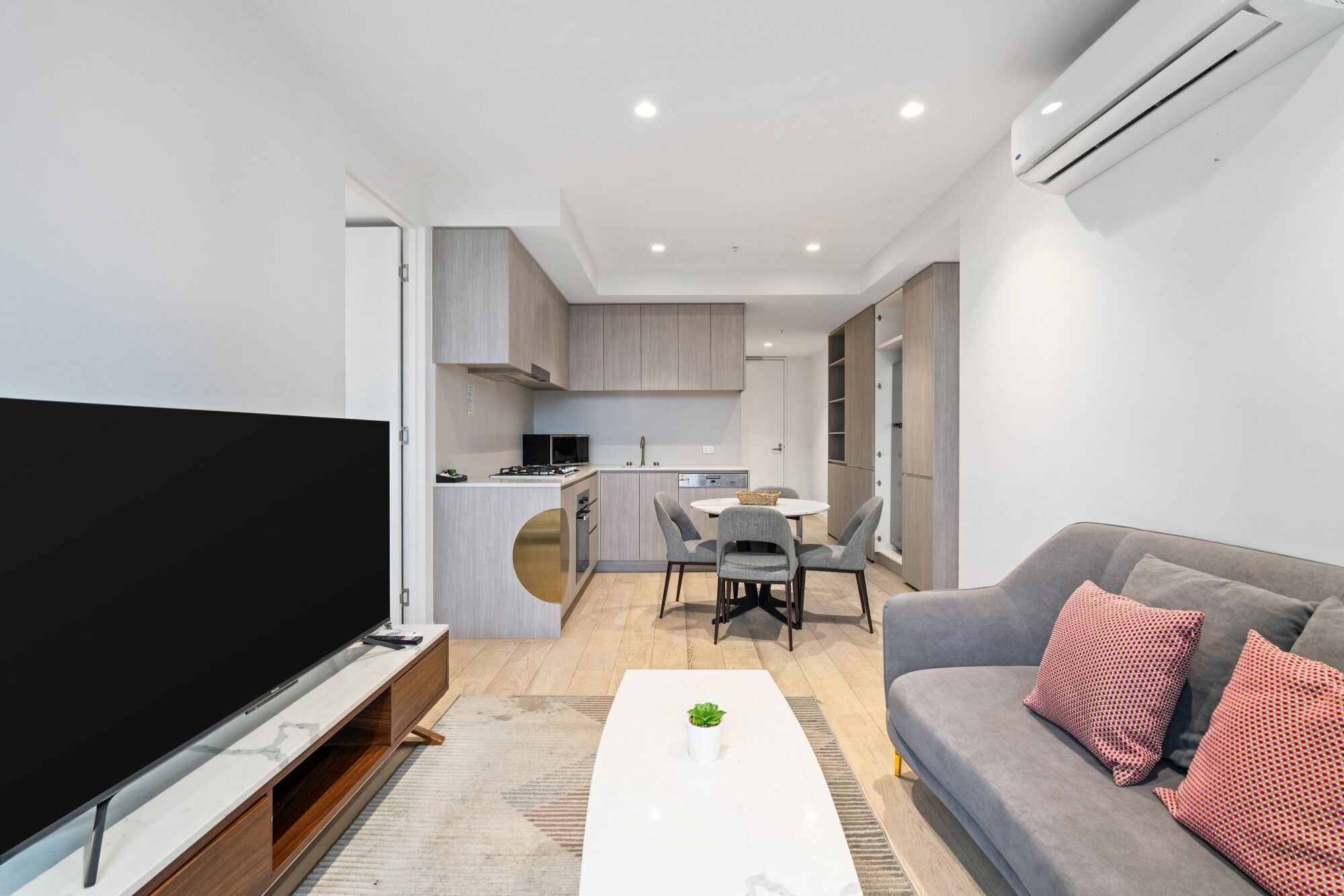Un soggiorno moderno con una televisione a schermo piatto, un divano grigio con cuscini decorati, un tavolino da caffè bianco e un angolo cottura con tavolo e sedie.