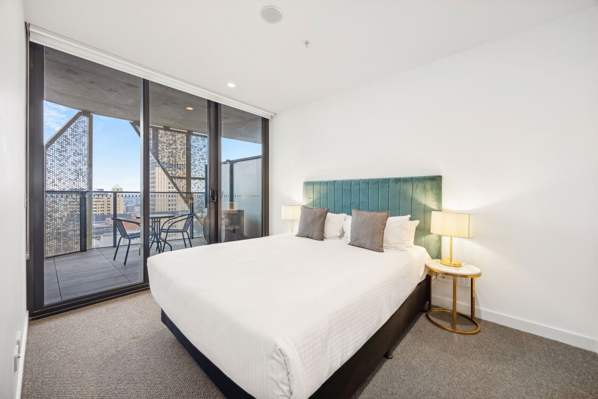 Een hotelkamer met een groot bed, een nachtkastje met lamp, een balkon met uitzicht op de stad en een schuifdeur van glas.