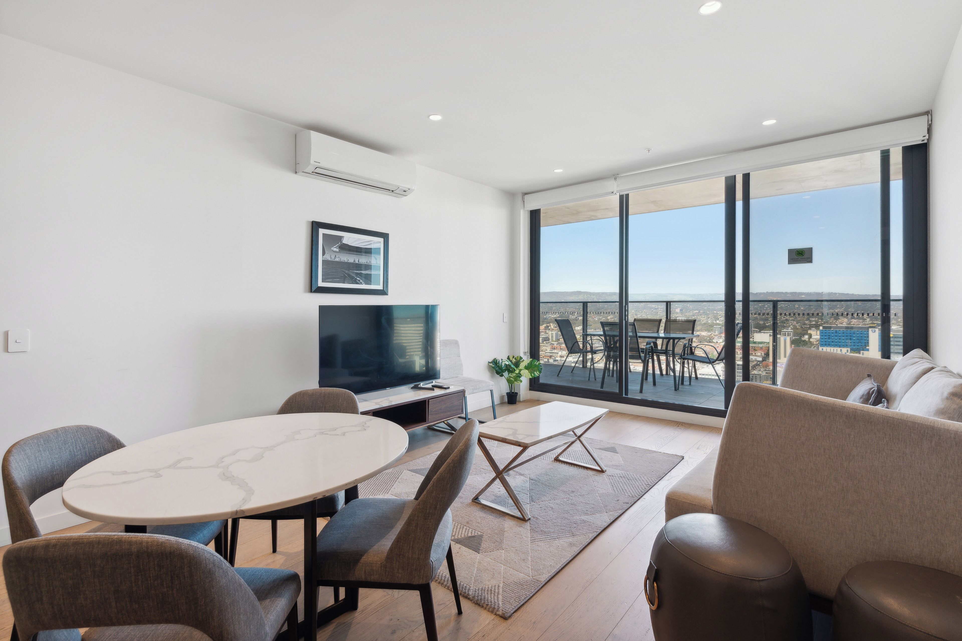 A modern living room with a dining table, chairs, a sofa, a TV, and a large window with a city view.