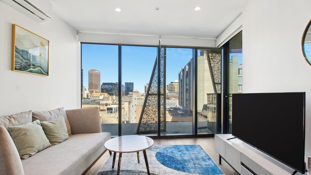 Una sala de estar moderna con un sofá, una mesita redonda, un televisor de pantalla plana grande y vistas a un paisaje urbano a través de amplios ventanales.