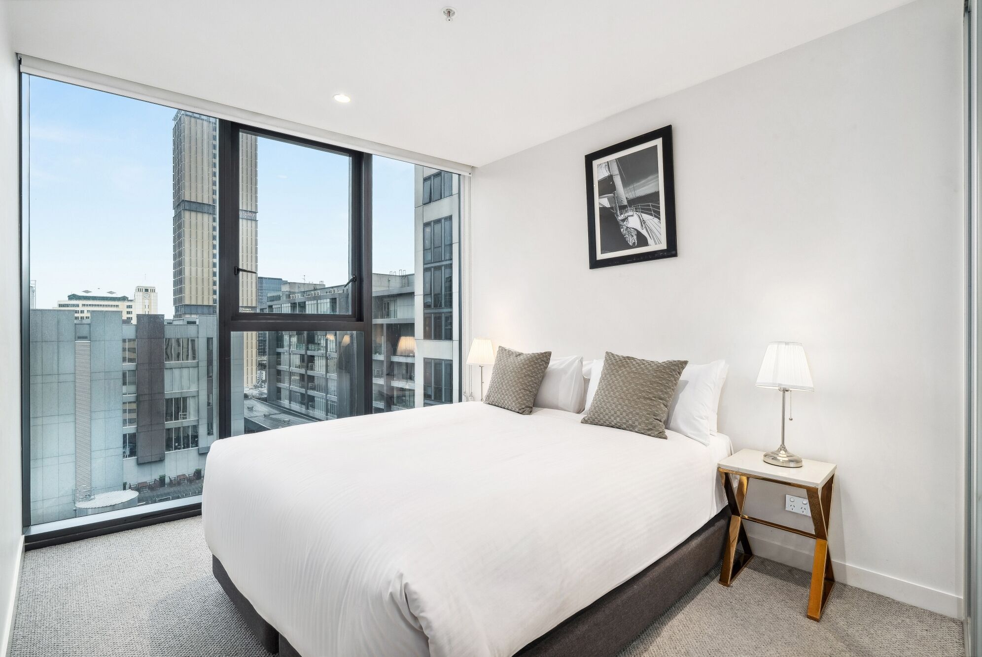 A modern hotel room with a large bed, a framed picture on the wall, and a view of the cityscape through the window.