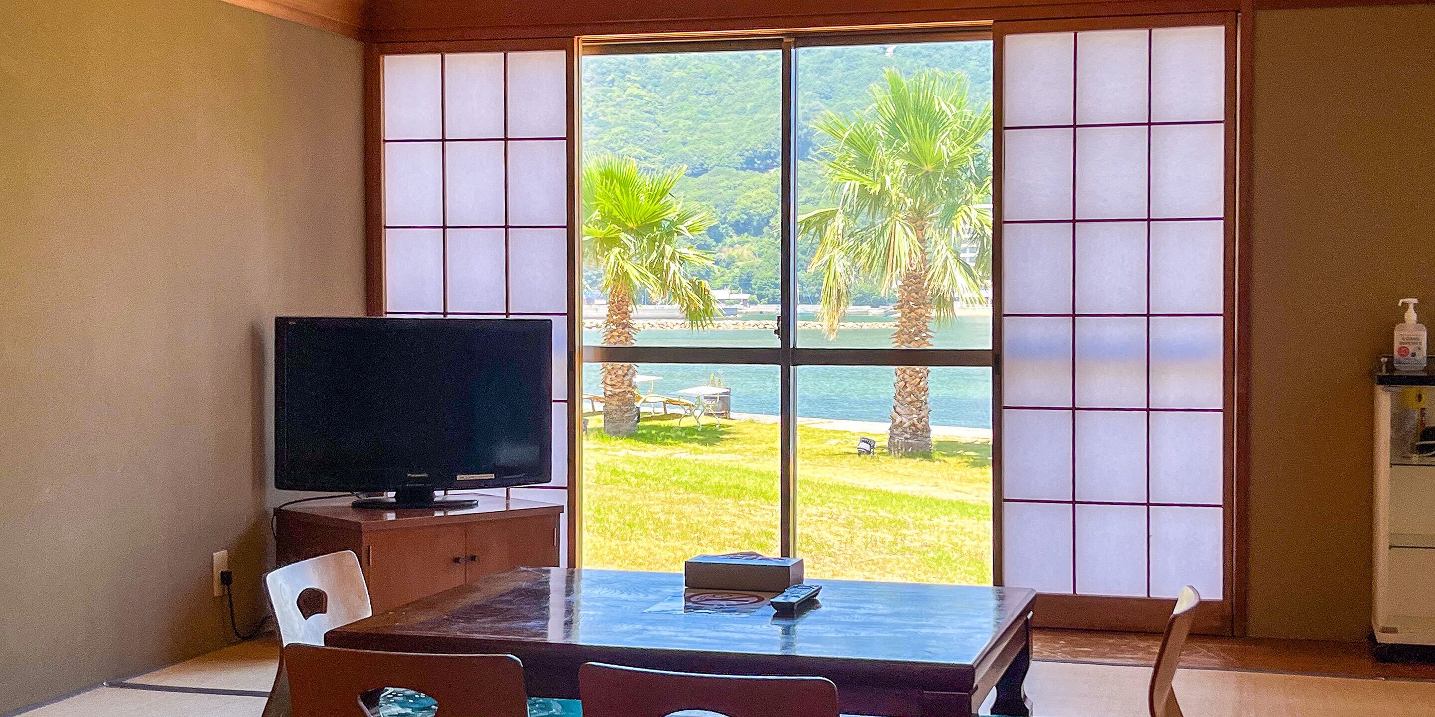 A room with a TV on a wooden stand, a table with chairs, and a view of palm trees and a beach through large shoji screens.