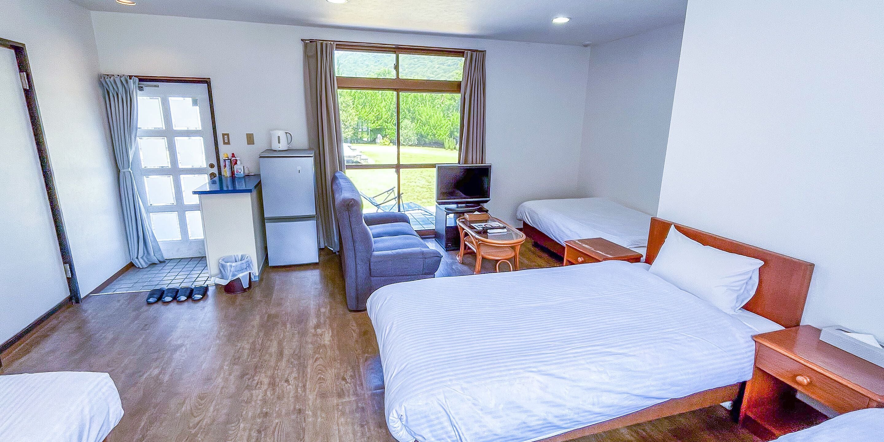 Habitación de hotel con una cama grande, una cama más pequeña, un sofá, un televisor, un escritorio y una ventana con vista a la vegetación.