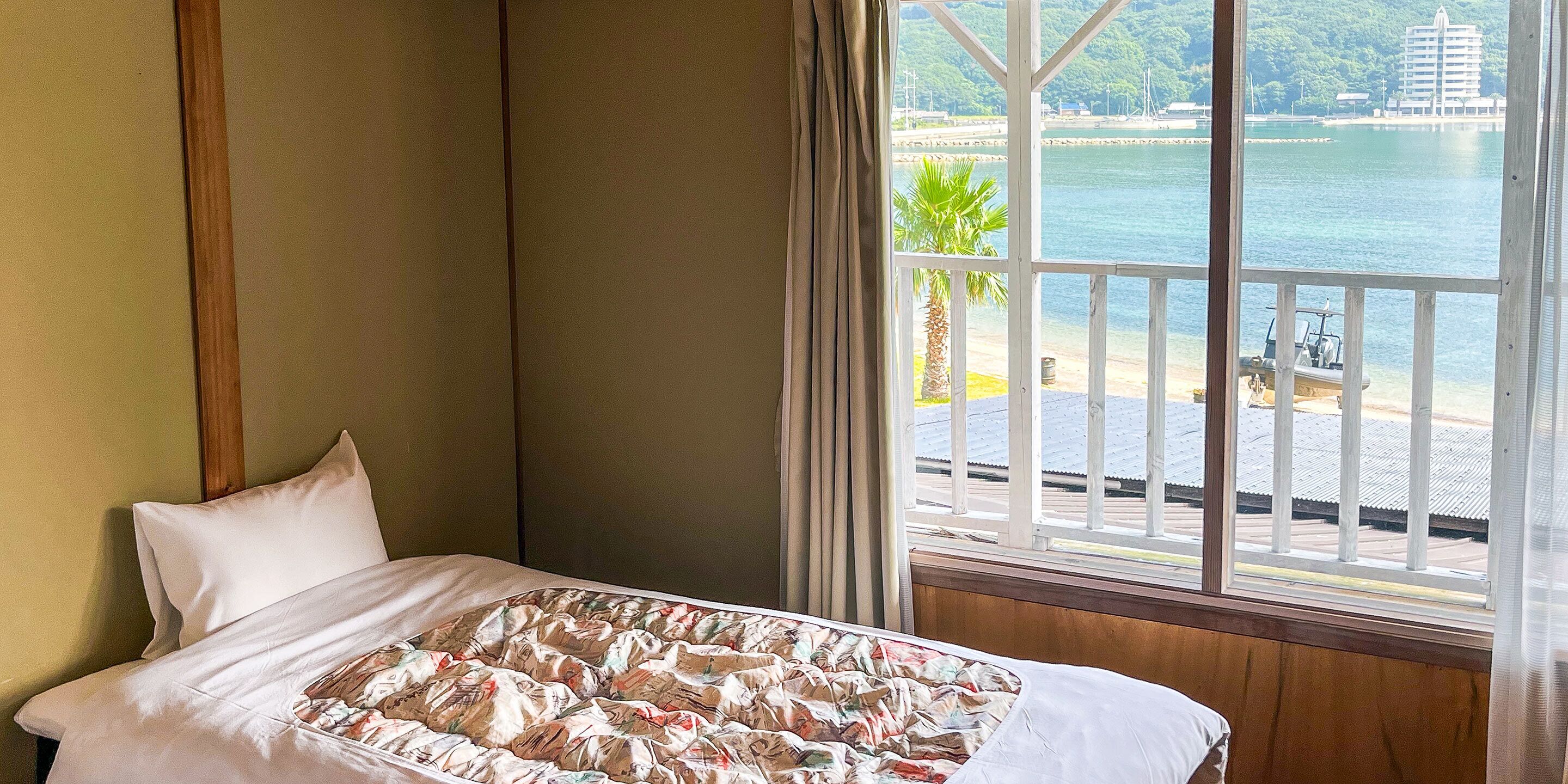 A hotel room with a bed, a window with a view of a beach and water, and a wooden floor.