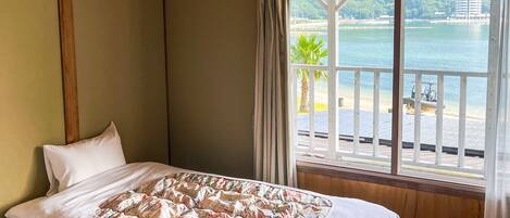A hotel room with a bed, a window with a view of a beach and water, and a wooden floor.