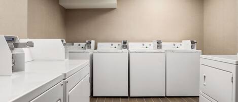 A laundry room with four white washers and dryers, a countertop with a paper towel dispenser, and a tiled floor.