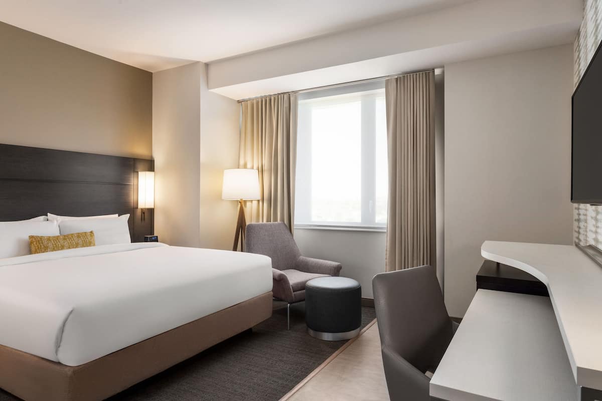A hotel room with a large bed, a desk, a chair, a lamp, and a window with curtains.