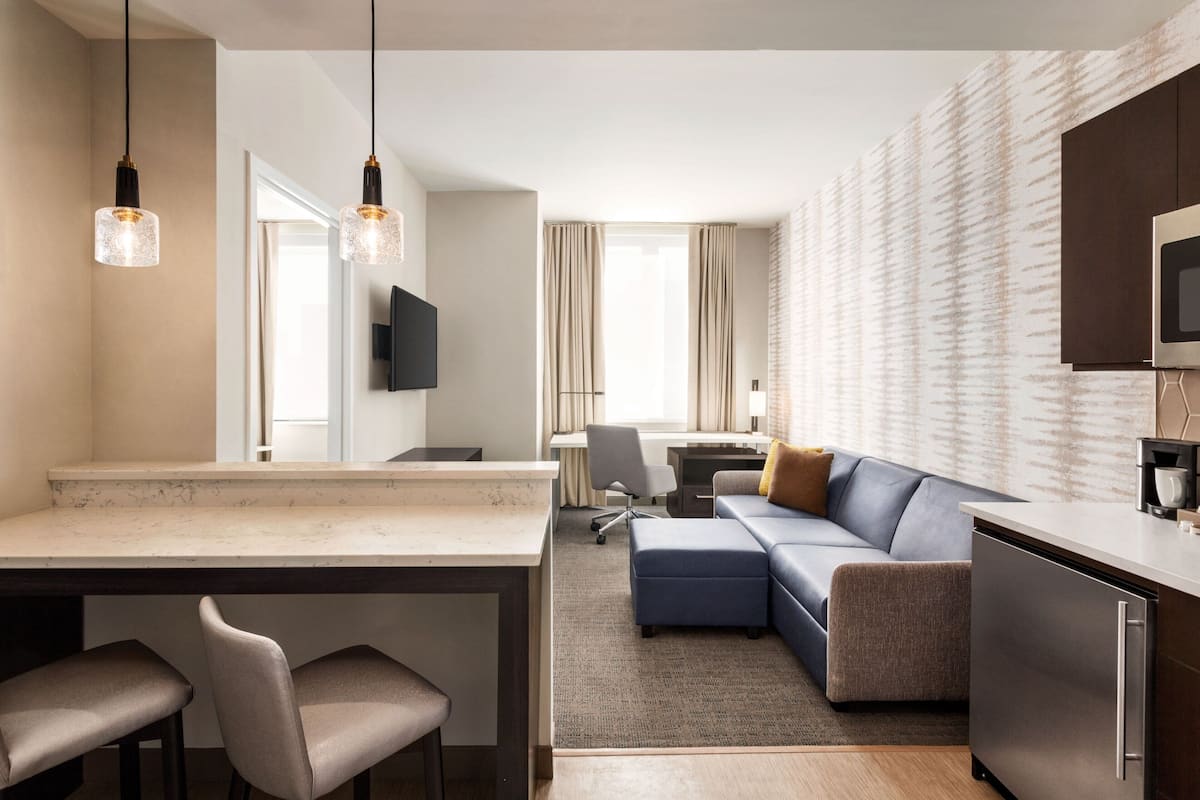 A modern hotel room with a kitchen area, a living space with a sofa and ottoman, and a dining area with a table and chairs.