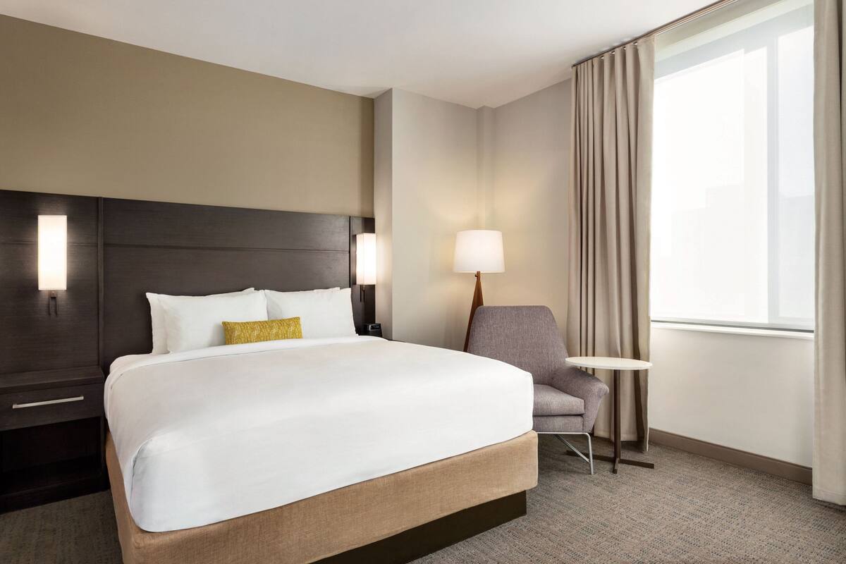 A hotel room with a large bed, a bedside table, a chair, a lamp, and a window with curtains.