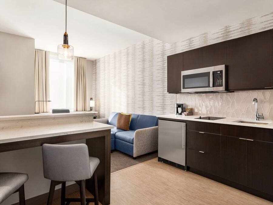 A modern kitchen with dark cabinets, a microwave, and a sink. There is a bar area with stools and a window with curtains.