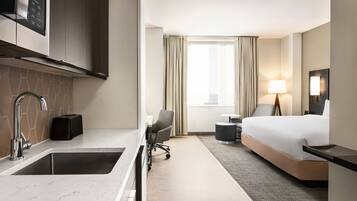 A modern hotel room with a kitchen area, a bed, a desk, and a window with curtains.