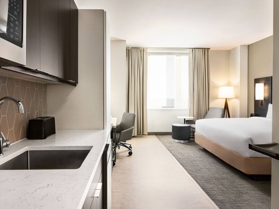 A modern hotel room with a kitchen area, a bed, a desk, and a window with curtains.