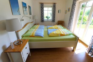 Quarto com uma cama de casal, mesa de cabeceira com luminária, janela com cortinas e vista para o verde lá fora.