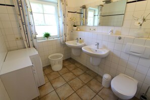Uma casa de banho com duas pias, sanita, janela com cortinas, uma planta num vaso e um espelho sobre as pias.