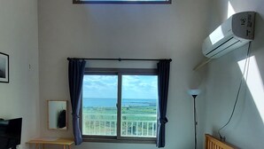 Quarto com uma cama grande, escrivaninha, cadeira, televisão, janela com cortinas e ar condicionado.