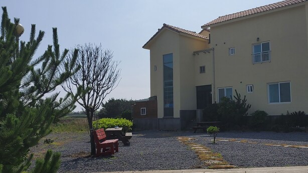 Um edifício de dois andares com telhado plano e grandes janelas. Uma entrada com calçamento de brita leva a um banco de madeira e uma pequena árvore.