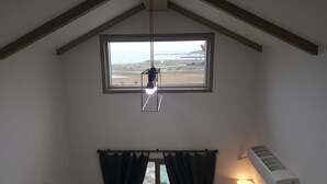 Quarto com teto inclinado, janela com vista para a paisagem, luminária pendente e ar-condicionado fixado na parede.