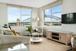 A modern living room with large windows, a flat-screen TV, a sofa, a coffee table, and a cabinet. - Harbourside 76 (McMahons Point)