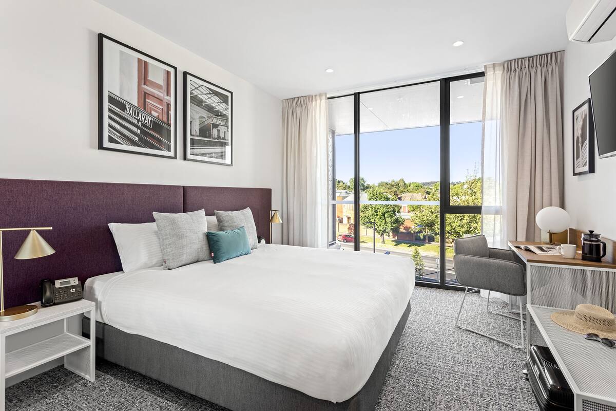 A modern hotel room with a large bed, a desk with a chair, a TV, and a view of the outdoors.