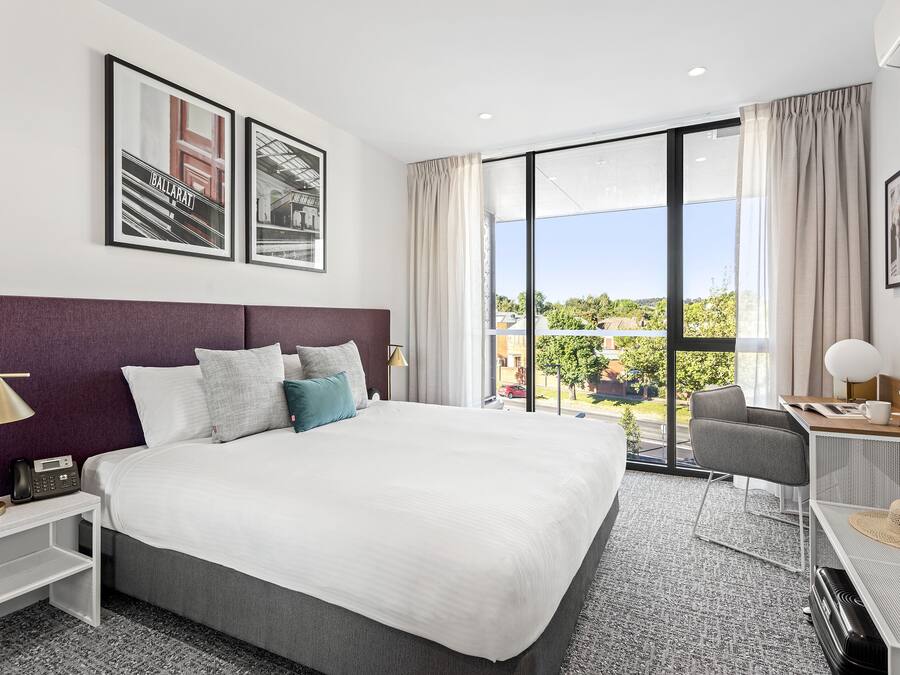A modern hotel room with a large bed, a desk with a chair, a TV, and a view of the outdoors.