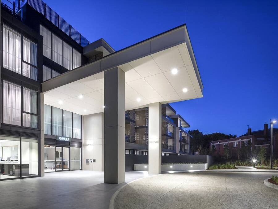 Une entrée d’hôtel moderne avec un espace extérieur abrité, de grandes fenêtres et un restaurant bien visible appelé « Quest ».