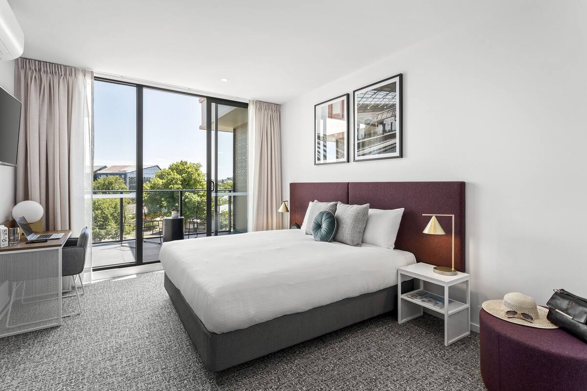 A modern hotel room with a large bed, a desk with a laptop, a small table with a lamp, and a balcony with a view of the city.