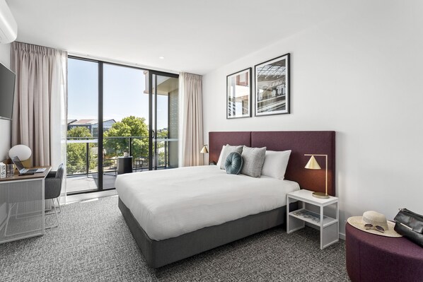 Una habitación de hotel moderna con una cama grande, un escritorio con una computadora portátil, una mesita con una lámpara y un balcón con vista a la ciudad.