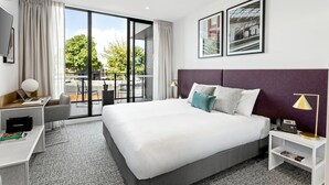 A modern hotel room with a large bed, a desk, a laptop, a chair, a TV, and a balcony with a view of greenery and a building. - Quest Ballarat Station (Soldiers Hill)
