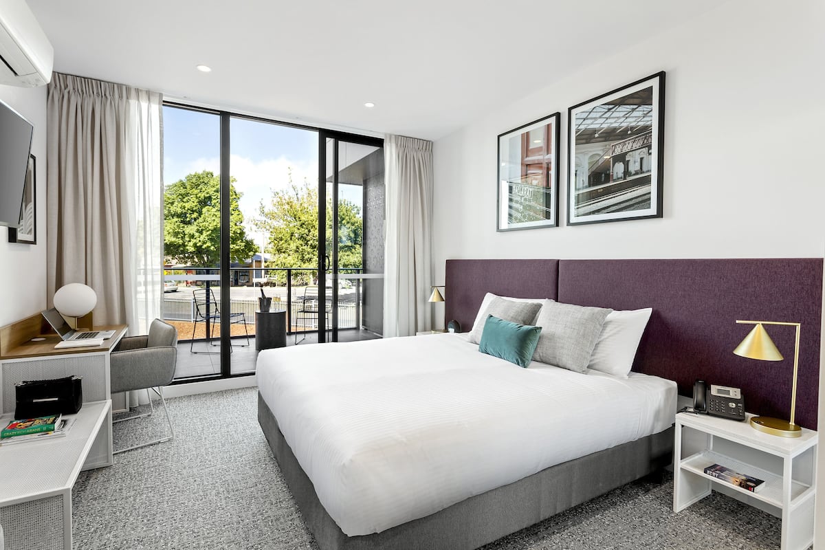 A modern hotel room with a large bed, a desk, a laptop, a chair, a TV, and a balcony with a view of greenery and a building.