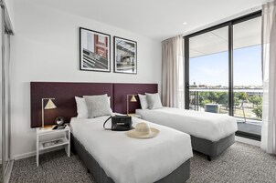 Habitación de hotel con dos camas, un ventanal con vistas a la ciudad, un cuadro en la pared y una mesita de noche con una lámpara y un bolso.