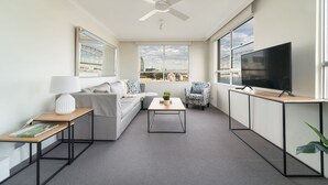 Una sala de estar moderna con sofá, mesa de centro y televisor, ventanas amplias con vista a la ciudad y un ventilador de techo.