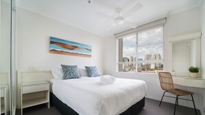 Una habitación de hotel con una cama grande, un escritorio, una silla, un ventanal con vista a la ciudad y un ventilador de techo.