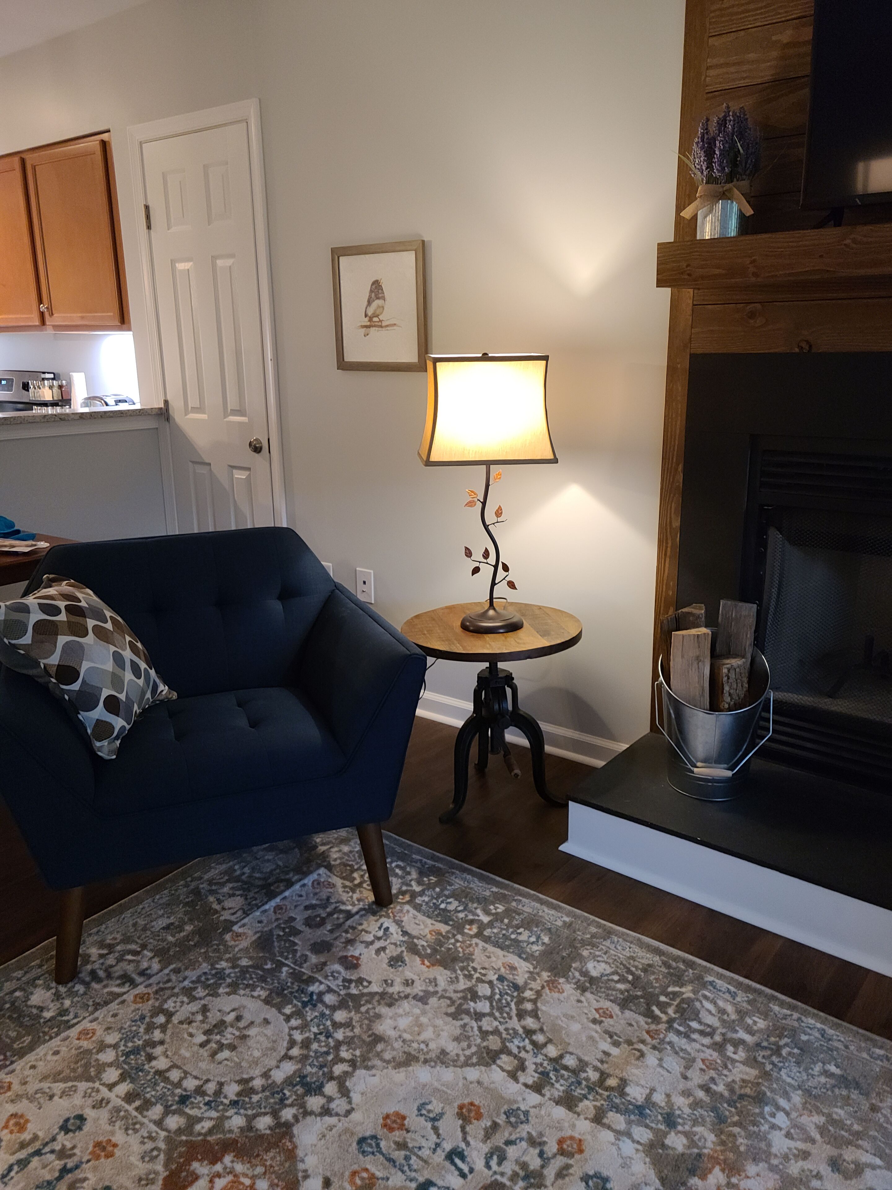 A living room with a blue armchair, a small round table with a lamp, a fireplace, and a kitchen in the background.